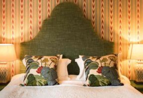 A cosy, country bedroom featuring a large bed with striking green headvoard and floral cushion ?>