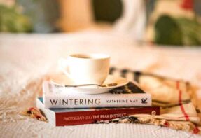 White cup of coffee and books stacked on white bed sheets ?>